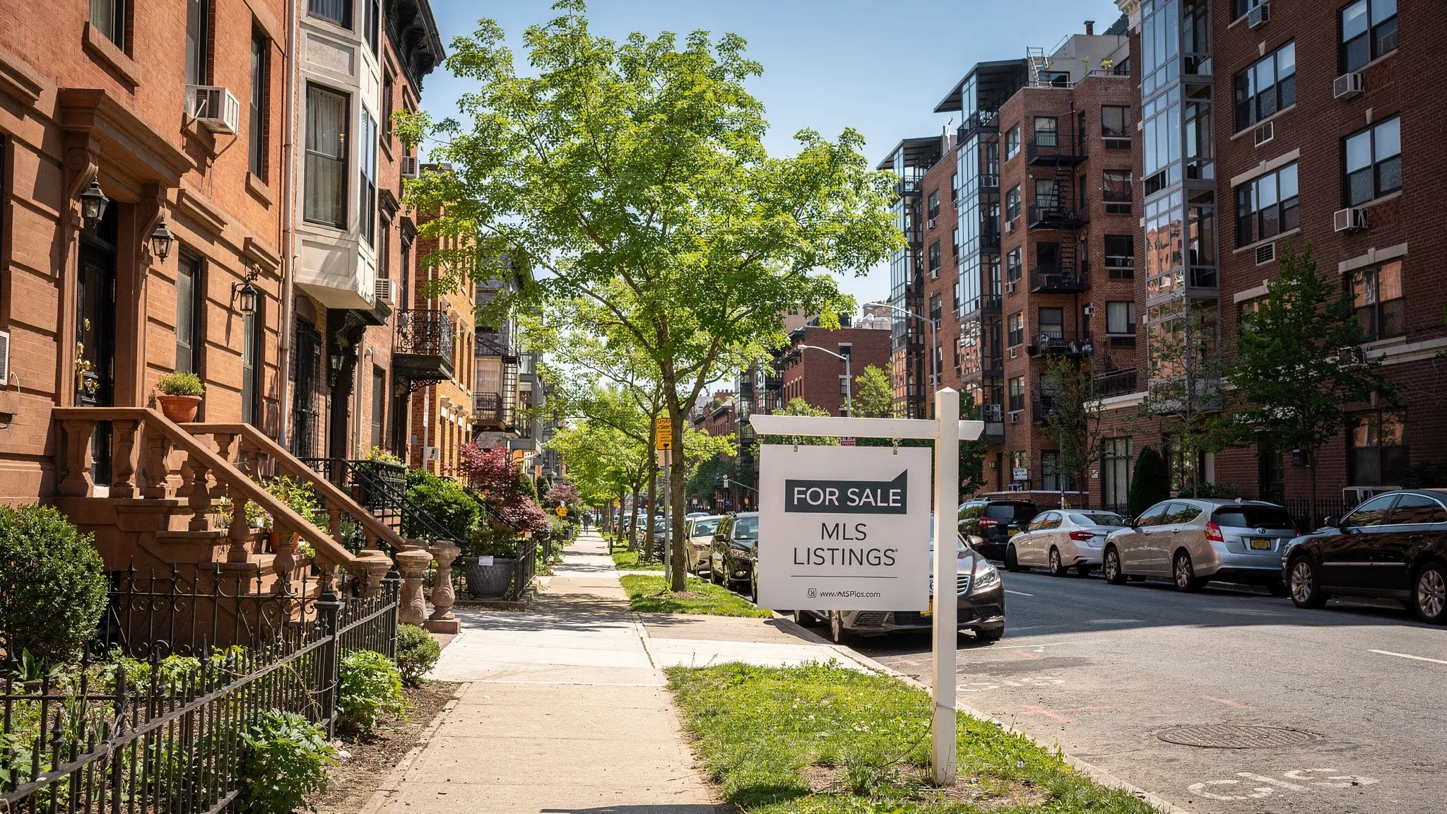 A New York residential street with townhomes, apartment buildings, and a real estate sign in the foreground, showing the variety of property types buyers may find through MLS listings.